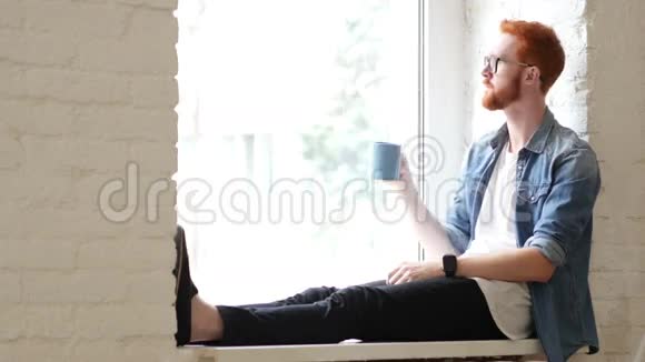 昂贵的设计师喝咖啡坐在窗口红色头发和胡子视频的预览图
