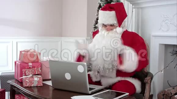 忙碌的圣诞老人用手提电脑和数码平板电脑准备礼物整理他的信件并收到一份来自视频的预览图