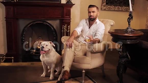 一个有钱人坐在房间里的椅子上壁炉和威士忌忠诚的狗躺在脚边视频的预览图
