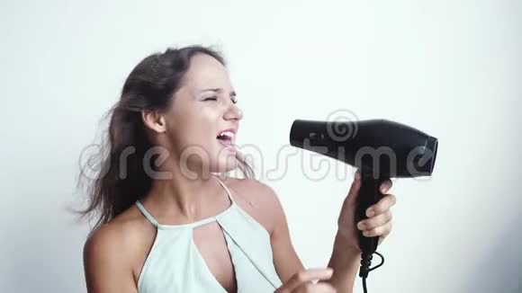 美丽的年轻快乐的女人在白色背景上使用吹风机唱歌慢动作3840x2160视频的预览图