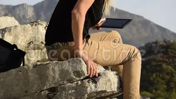年轻的金发女子坐在山崖边用数码平板电脑视频的预览图