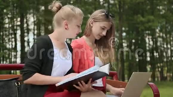 两个年轻漂亮的女学生手拿笔记本电脑在绿色公园的长凳上学习讨论侧视视频的预览图