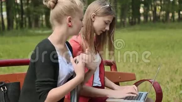 两个年轻漂亮的女学生手拿笔记本电脑在绿色公园的长凳上学习讨论侧视视频的预览图
