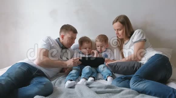 带平板电脑的家人妈妈爸爸和两个儿子双胞胎蹒跚学步的孩子看着躺在床上的平板电脑视频的预览图