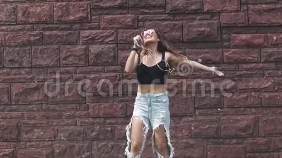 一个湿漉漉的女孩用手机而不是麦克风唱歌在街上的浪花下疯狂地跳舞视频的预览图
