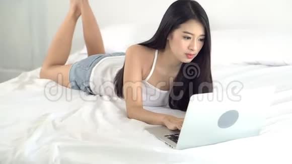 美丽的亚洲年轻女子躺在床上用笔记本电脑在卧室里休闲放松成功视频的预览图