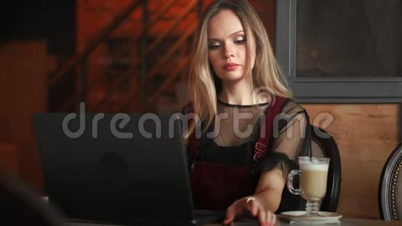 年轻女子坐在木桌上的咖啡店里喝咖啡用智能手机桌子上有笔记本电脑女孩视频的预览图