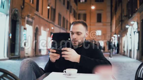 年轻人晚上坐在城市咖啡馆里拿着智能手机迷人的男性独自喝咖啡视频的预览图