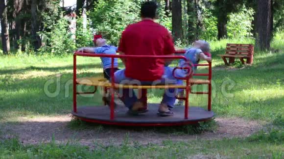 父亲带着两个孩子在夏天的绿色公园乘坐旋转木马视频的预览图