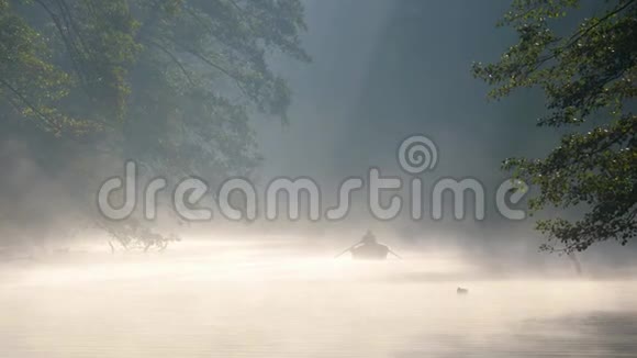 孤独的人带着桨在船上在黎明的雾中平静的河流上游泳清晨的第一缕阳光使他们视频的预览图