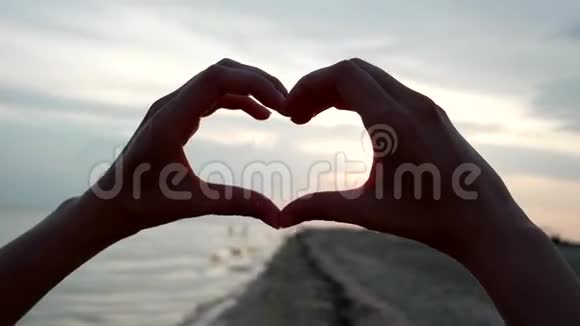 心手在海滩上的日落时分展现太阳透过一个爱的标志看着镜头视频的预览图