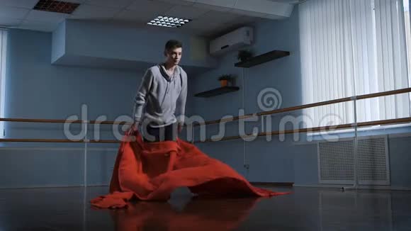 身穿灰色套头衫和黑色长裤的帅气男舞者正在教室中央用红布跳舞年轻的视频的预览图
