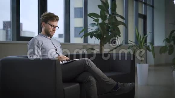 穿着蓝色衬衫的留着胡子的员工正坐在办公室的沙发上用平板电脑视频的预览图