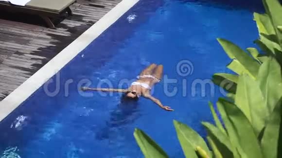 一个穿着白色泳衣的瘦女孩在游泳池里背上漂浮美丽的年轻女子在度假视频的预览图
