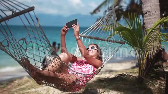年轻可爱的女孩戴着太阳镜躺在户外的秋千床上享受日光浴用手机自拍视频的预览图