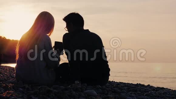 坐在鹅卵石海滩上享受石碑的男女剪影日落时大海或湖泊后视图视频的预览图