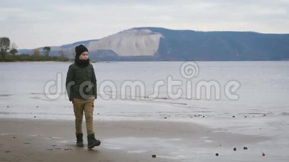 穿着深灰色夹克芥末裤黑色帽子和围巾的时髦少年独自沿着海滨海岸散步视频的预览图
