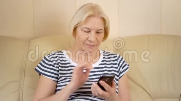 开朗好看的高级女性坐在家里的沙发上在她的智能手机上聊天视频的预览图