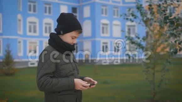 穿着暖色灰色夹克戴着黑色帽子和围巾的孩子正在大街上打电话街上有许多建筑视频的预览图