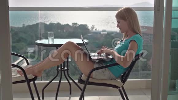 美丽迷人的女人坐在阳台的椅子上浏览互联网在笔记本电脑上阅读新闻视频的预览图