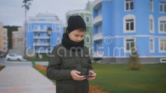 穿着温暖的深灰色夹克和黑色帽子和围巾的孩子站在街上玩电话男孩正在给视频的预览图