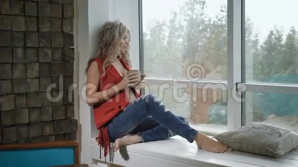 年轻的孕妇穿着红色衬衫端着一杯茶坐在窗台上美丽的未来母亲喝茶视频的预览图