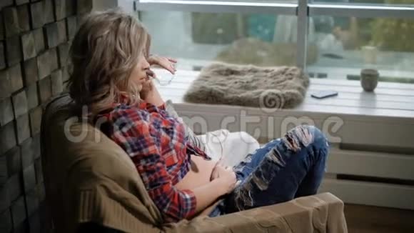 年轻漂亮的孕妇穿着格子衬衫和蓝色牛仔裤坐在靠近窗户的扶手椅上她在吹她的头发视频的预览图