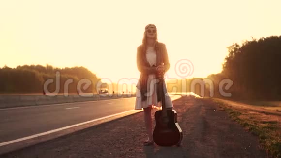 带吉他的女时髦女郎在日落的路上旅行戴太阳镜的年轻漂亮女孩的肖像女孩视频的预览图