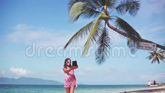 美丽的黑发女郎穿着红色连衣裙戴着太阳镜在棕榈树下的沙滩上用手机自拍视频的预览图