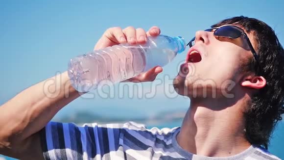 健康的年轻人戴着太阳镜从瓶子里喝水在海景背景下慢动作1920x1080视频的预览图