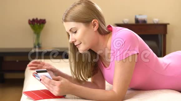 美丽迷人的年轻女子躺在沙发上用信用卡在黑色平板电脑上在线购买视频的预览图