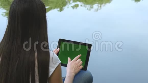 年轻女孩使用数字平板电脑和绿色屏幕坐在湖边的木制码头后视图视频的预览图