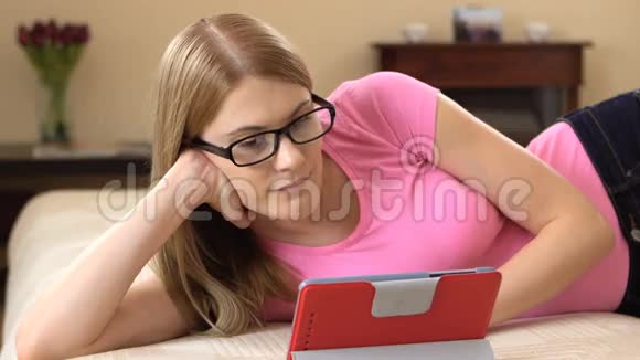 漂亮迷人的年轻女人戴着眼镜平板电脑躺在沙发上浏览互联网微笑视频的预览图