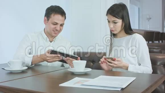 微笑的年轻人在他的平板电脑上显示一些东西给女同事在午餐时拿着电话视频的预览图