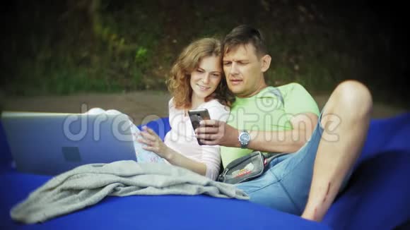年轻女子和成熟男子使用笔记本电脑躺在公园的休息室里轻轻地吸着烟视频的预览图