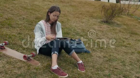 年轻的女学生带着背包和滑板躺在草地上带着书在户外公园做作业视频的预览图
