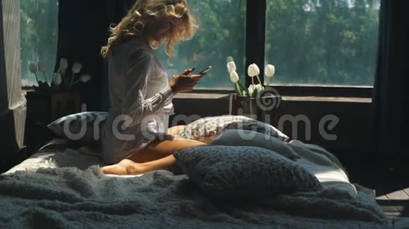 年轻漂亮的金发女郎正坐在窗边的床上在电话上打印一条信息视频的预览图