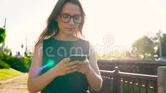 戴眼镜的女人在日落时分走在街上时使用智能手机视频的预览图