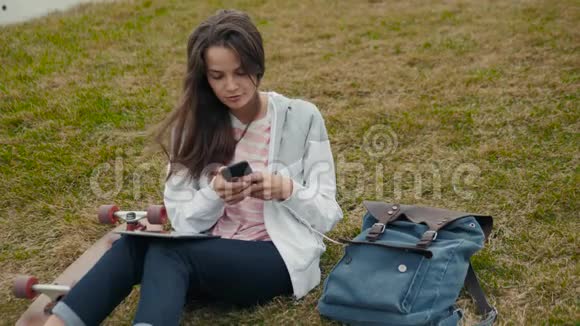 年轻的女学生带着背包和滑板躺在草地上带着书在户外公园做作业视频的预览图