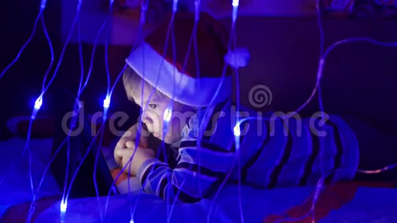 孩子看着躺在床上戴着帽子的药片圣诞老人前景上灯光花环视频的预览图