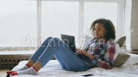 快乐的混血女孩躺在床上和朋友用笔记本电脑聊天视频的预览图