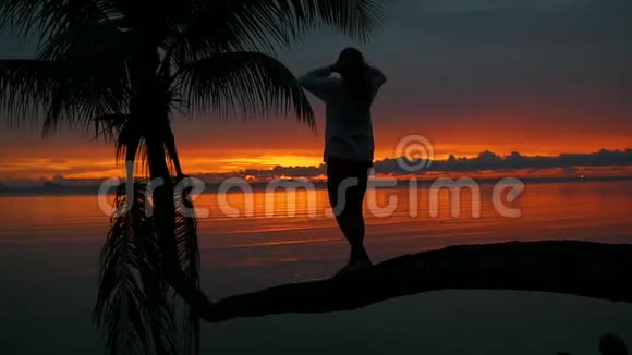 一个女孩站在海滩上红日落的背景下的棕榈树上视频的预览图