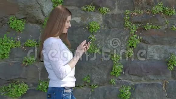 漂亮的年轻女孩走在老式的野生石头墙和使用智能手机视频的预览图