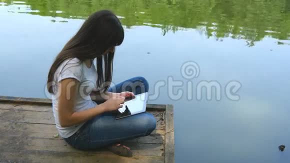 女孩使用平板电脑在湖边放松坐在靠近水面的木制码头的边缘视频的预览图