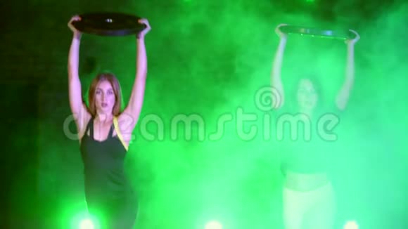 两个运动型的漂亮的女人用厚重的盘子做力量练习晚上在轻烟雾中视频的预览图