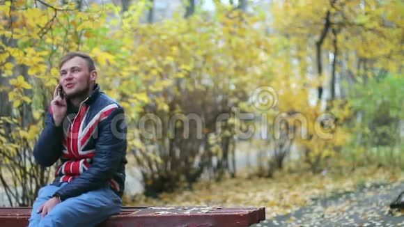 美丽快乐的年轻人坐在黄色灌木丛附近的长凳上用手机说话视频的预览图