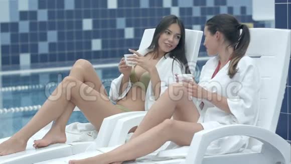 两位微笑的年轻女士坐在泳池边的躺椅上喝茶视频的预览图