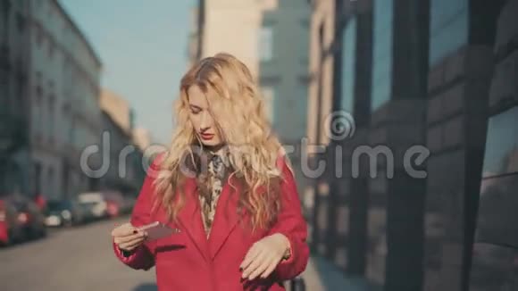 漂亮的年轻女子穿着红色外套在坚硬的阳光下使用智能手机走向阴影稳定的凸轮射击视频的预览图