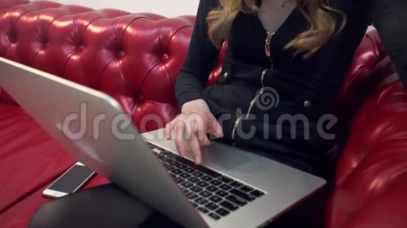 漂亮的金发美女在红色沙发上使用笔记本电脑并在客厅吃一个绿色苹果视频的预览图