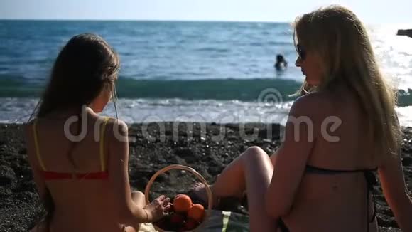 妈妈和女儿穿着泳衣留着长发在海浪背景下的海边吃水果和浆果视频的预览图